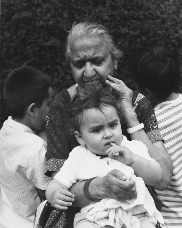 An elderly woman holds a baby on her lap, gently touching her face. Two other children, seen from behind, lean against her. They are sitting outdoors in front of dense bushes.