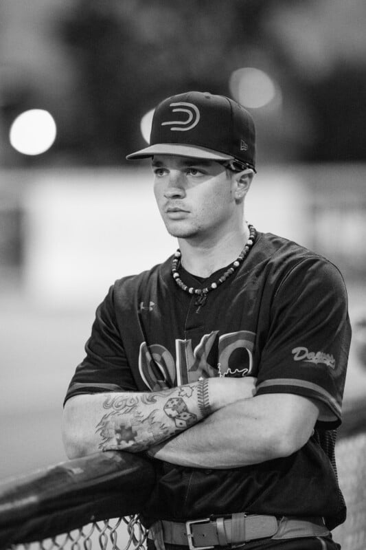 A baseball player in uniform with tattoos on his forearm stands with arms crossed, leaning on a fence. He wears a cap and a beaded necklace, looking focused. The image is black and white.