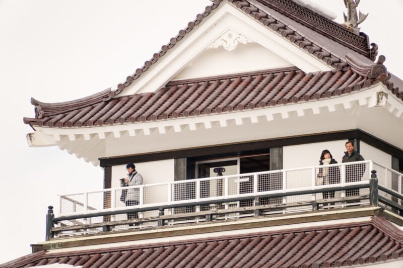 Three people stand on the balcony of a traditional Japanese-style building with ornate brown roof tiles. They appear to be observing the view, with one person holding a phone. The sky is overcast.