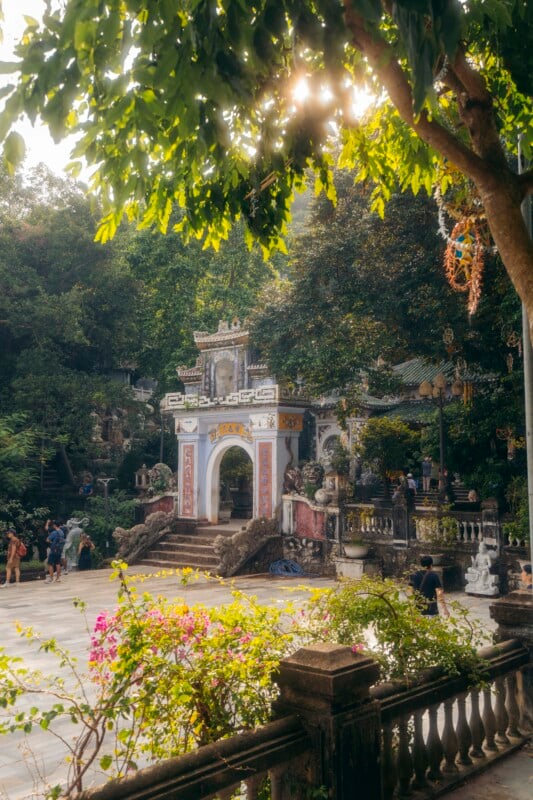 A sunlit temple gate with ornate decorations stands amid lush greenery and flowering plants, with people walking around in the peaceful courtyard. Sun rays filter through tree branches overhead.