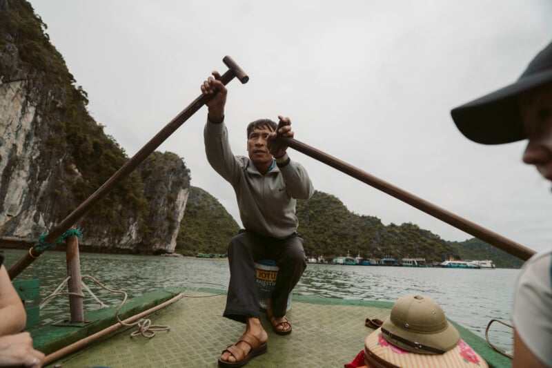 A man in a gray sweater rows a boat with wooden paddles on a greenish lake, surrounded by rocky cliffs and lush hills. Two people sit in the foreground, one wearing a hat. The sky is overcast.