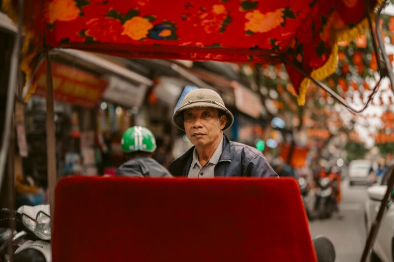 A man wearing a helmet and jacket stands on a busy street, partially shaded by a red patterned canopy. Shops, motorbikes, and another person in a green helmet are visible in the background.