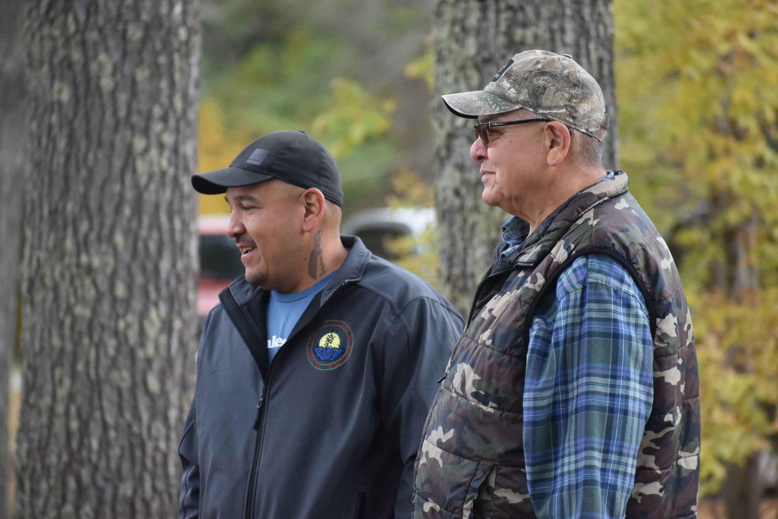 Two men stand outdoors among trees, both wearing jackets and caps; one has a camouflage vest and glasses, the other has a logo on his jacket.