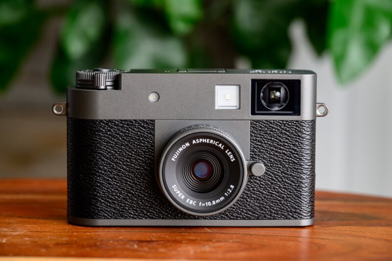 A black Fujinon rangefinder camera with a textured grip sits on a wooden surface, with green leaves blurred in the background.