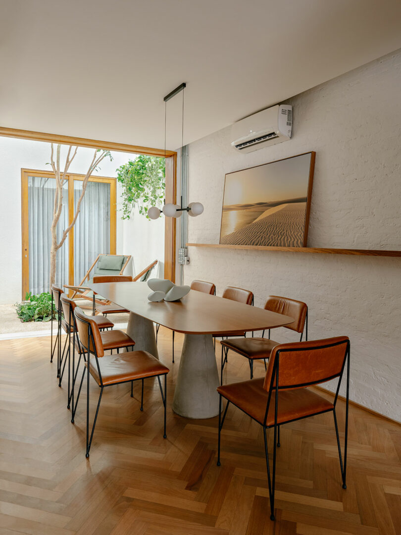 A modern dining room with a wooden table, six leather chairs, minimalist light fixture, wall art, indoor plants, and wood flooring, with natural light coming through a glass door.