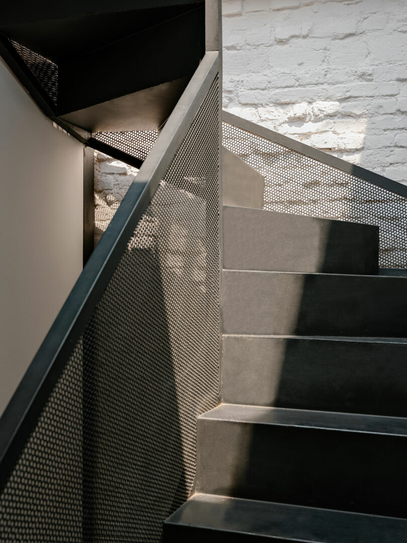 Black metal staircase with perforated railing and sunlight casting shadows against a white brick wall.