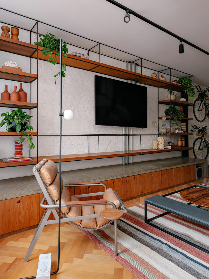 Modern living room with open shelving, a wall-mounted TV, indoor plants, a leather chair, striped rug, and a bicycle mounted on the back wall.