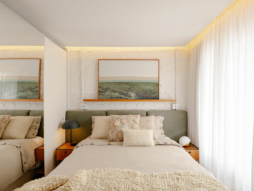 A neatly made bed with beige bedding and decorative pillows sits against a white brick wall, with a landscape photograph and bedside tables on each side. Sheer curtains let in natural light.