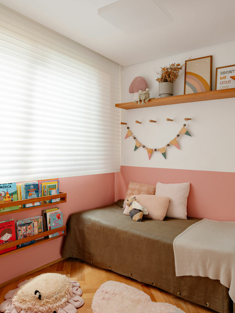 A cozy children's room with a bed, soft toys, shelves of books, pastel decorations, and a window with white blinds.