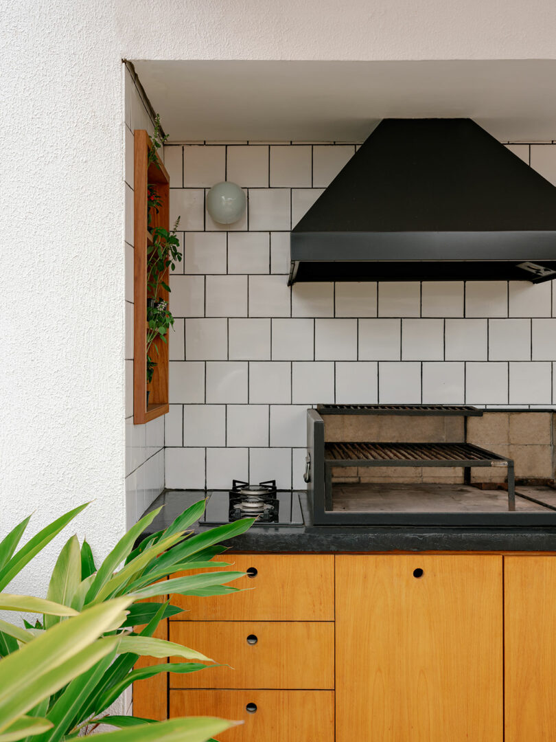 Outdoor kitchen with wood cabinetry, black countertop, a built-in grill, white subway tile backsplash, and a black range hood; green plants in the foreground and on the wall shelf.