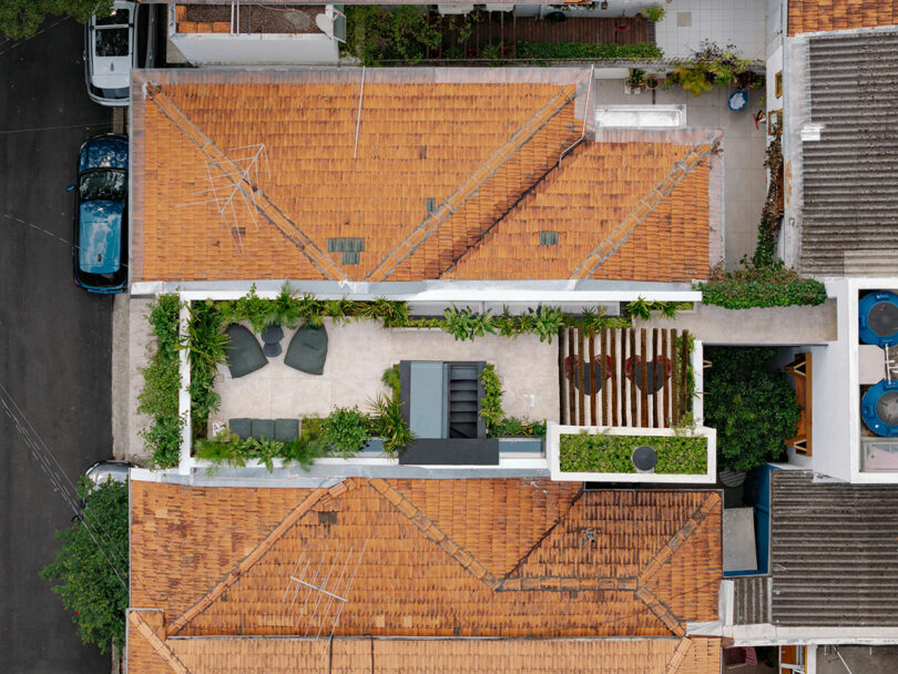 Aerial view of a narrow urban rooftop terrace with outdoor seating, greenery, and wooden slats, surrounded by orange-tiled roofs and adjacent buildings.