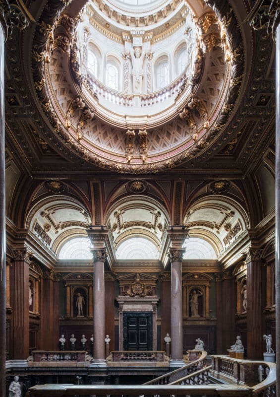 Ornate interior of a grand hall with a domed ceiling, intricate gold detailing, tall arches, marble columns, statues, and large windows letting in natural light.