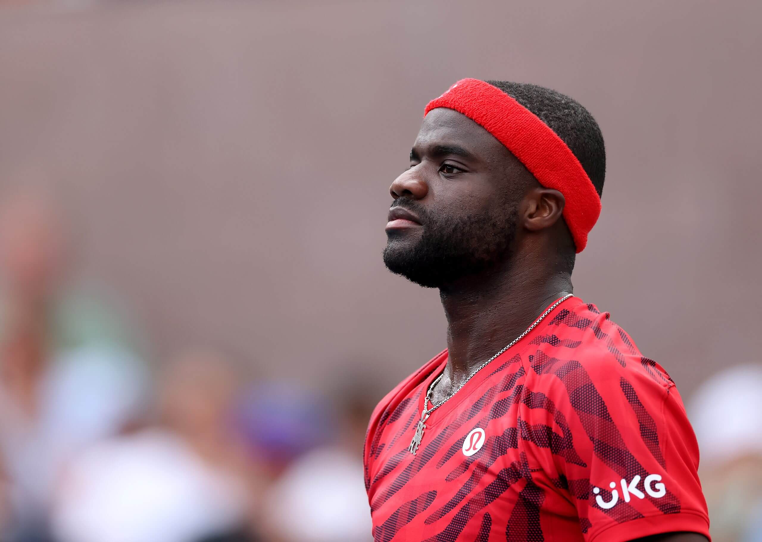 Frances Tiafoe looks on wearing a red headband and striped red v-neck shirt.