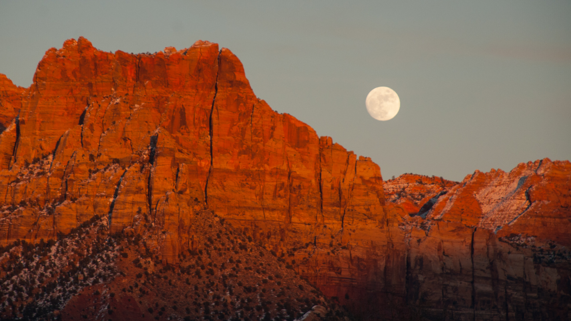 A full moon is seen in a gray sky over a series of red jagged cliffs.