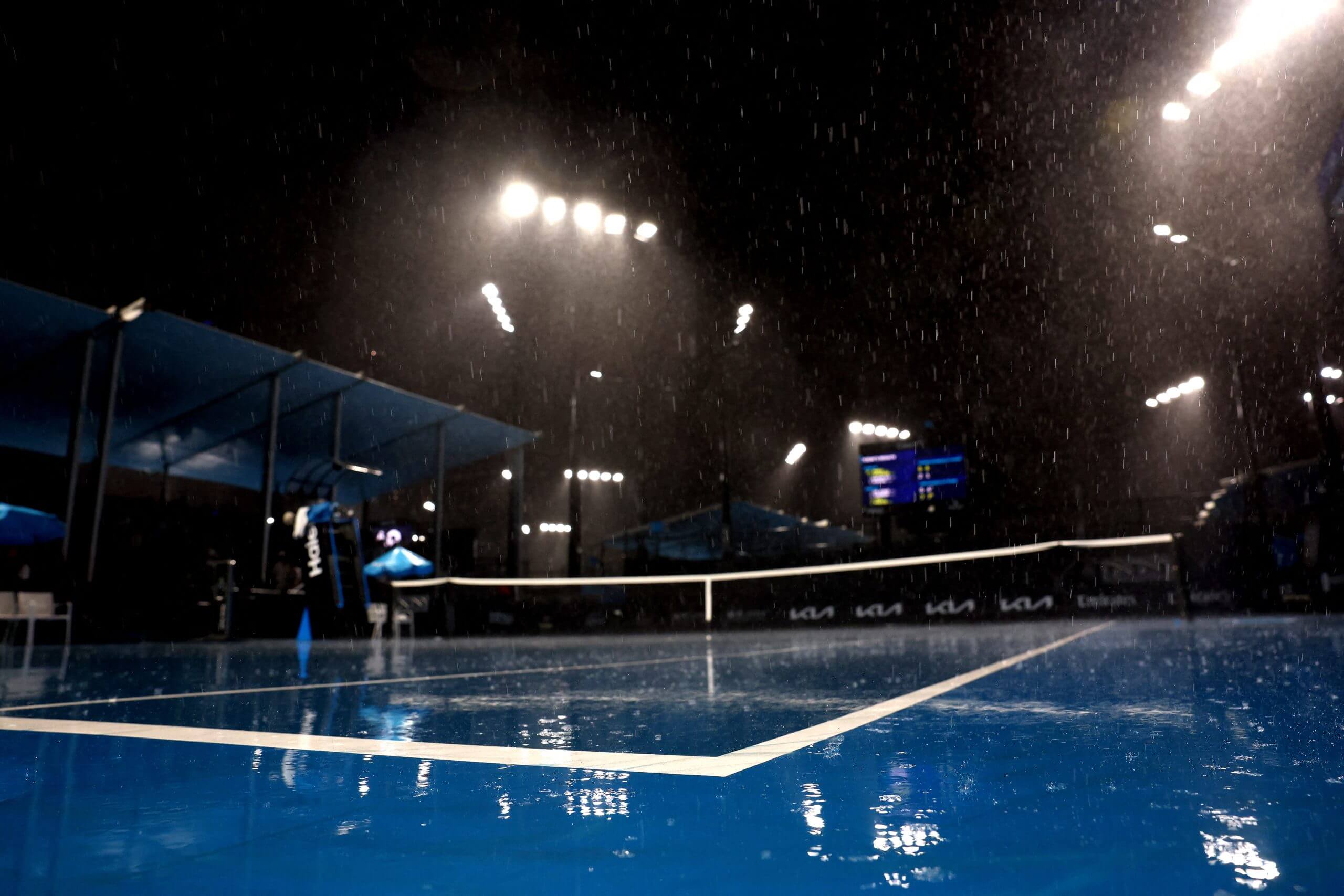 Rain falls against floodlights on a blue tennis court at night.