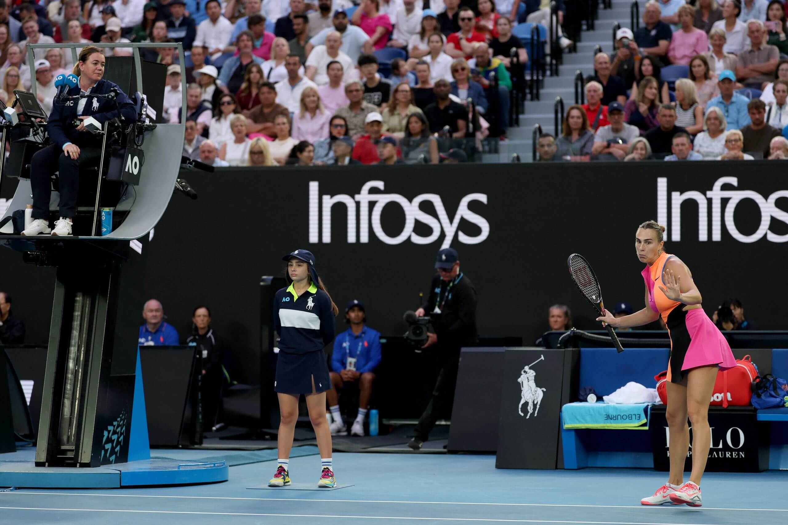 Aryna Sabalenka looks on wearing a multicolored tennis outfit while a chair umpire sits high to her left.