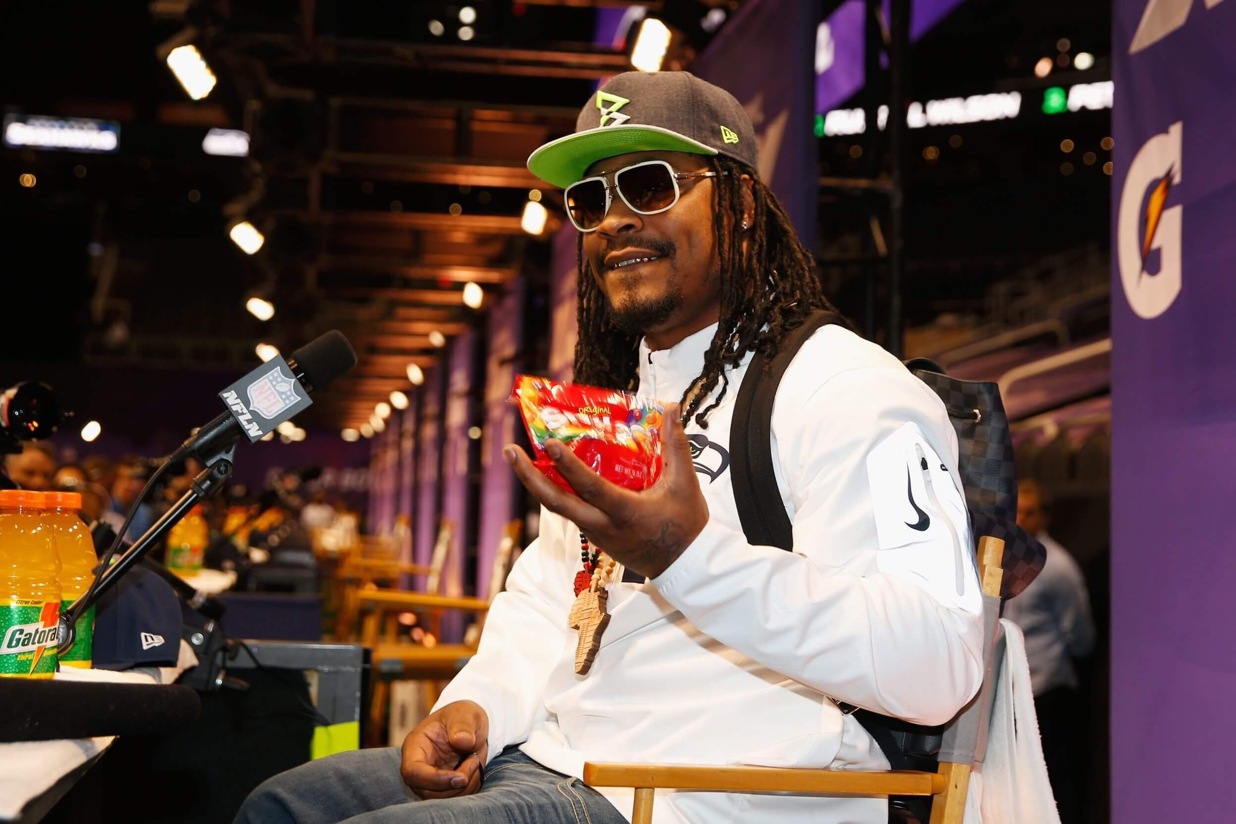 Marshawn Lynch, in a black hat with a neon green bill and white-rimmed sunglasses, eats Skittles in front of a microphone.