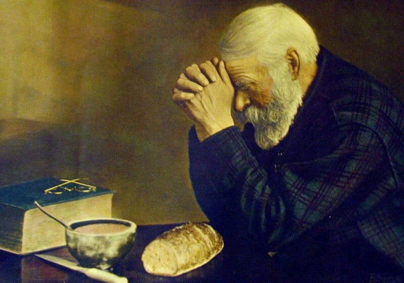 An elderly man with white hair and beard sits at a table, hands clasped in prayer, beside a bowl, a loaf of bread, a knife, and a book with a cross on its cover. The scene is warmly lit and contemplative.