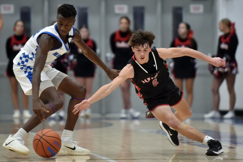 A basketball player in a white and blue uniform dribbles as a player in a black and red uniform lunges to steal the ball on a gym court, with cheerleaders blurred in the background.