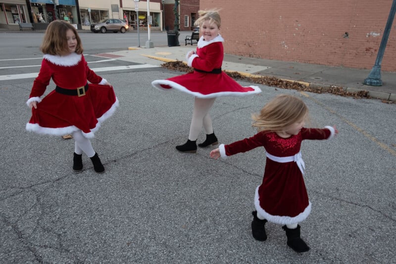 Three young girls in red holiday dresses twirl and play in an empty parking lot. The girls wear white tights and black boots, and two of the dresses have white fur trim. Buildings and shops are visible in the background.