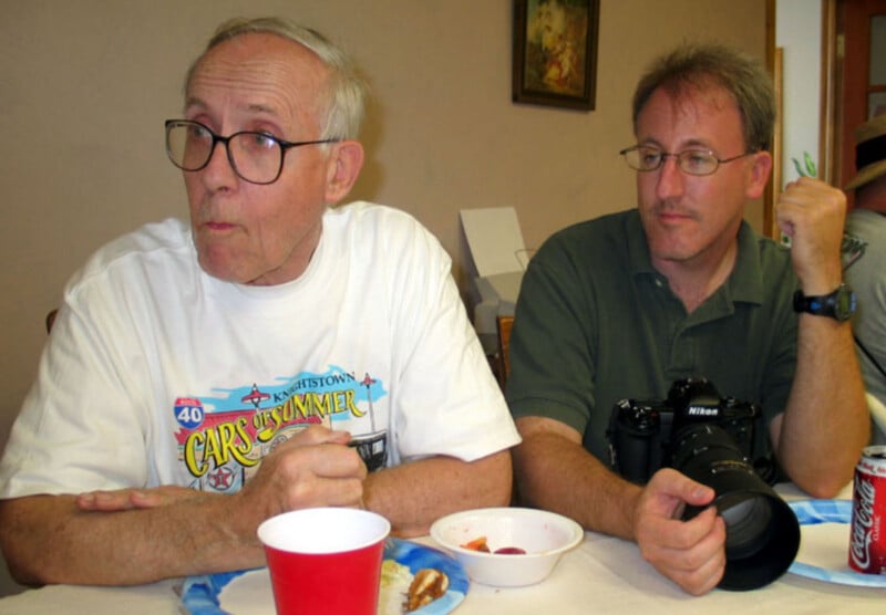 Two men sit at a table with food and drinks. The older man on the left wears glasses and a white "Cars of Summer" shirt. The younger man on the right has glasses, a camera, and a can of Coke in front of him.