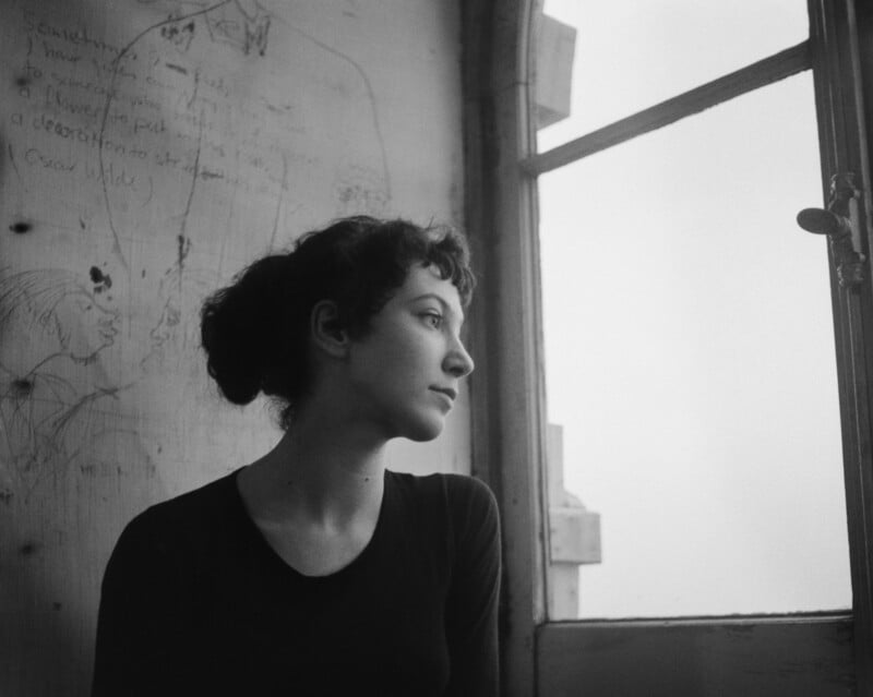 A woman with short, curly hair sits by a window, gazing outside. The wall behind her has sketches and handwritten notes. Soft, natural light fills the room, creating a calm, contemplative mood.