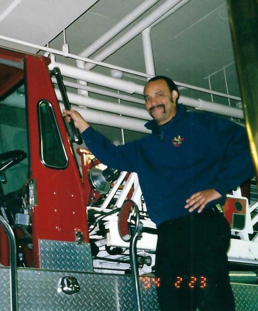 A person stands beside a red fire truck inside a building, holding onto the vehicle's door handle and smiling at the camera.