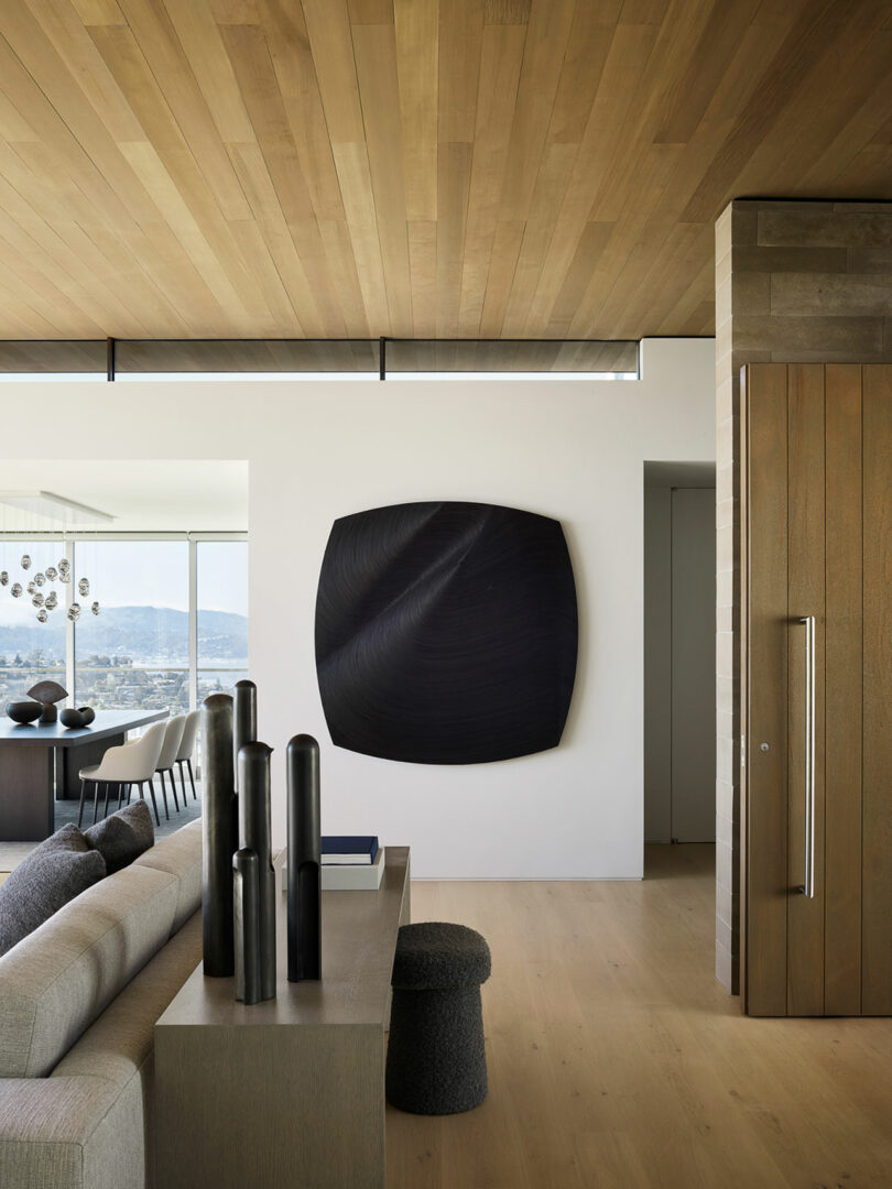 Modern living room with light wood floors and ceiling, a gray couch, black decorative vases on a console, and a large dark abstract wall art on a white wall.