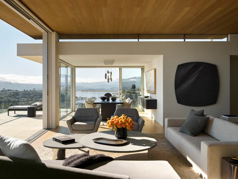 Modern living room with large glass doors open to a balcony, neutral furniture, a vase of orange flowers, and a view of hills and water in the background.