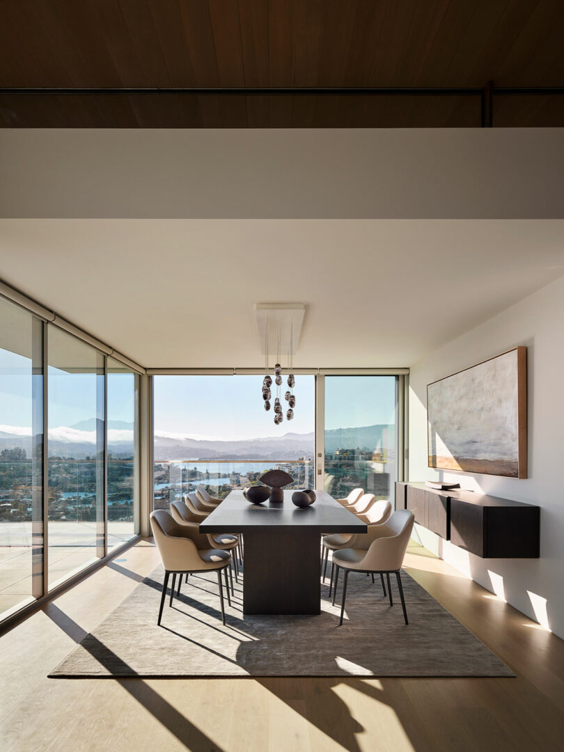 Modern dining room with a long table, ten chairs, large windows with city and mountain views, minimalist decor, and a contemporary chandelier.