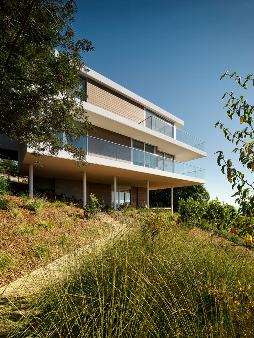 Three-story modern house with large glass balconies, elevated on stilts, surrounded by grasses and trees on a sloped landscape under a clear blue sky.