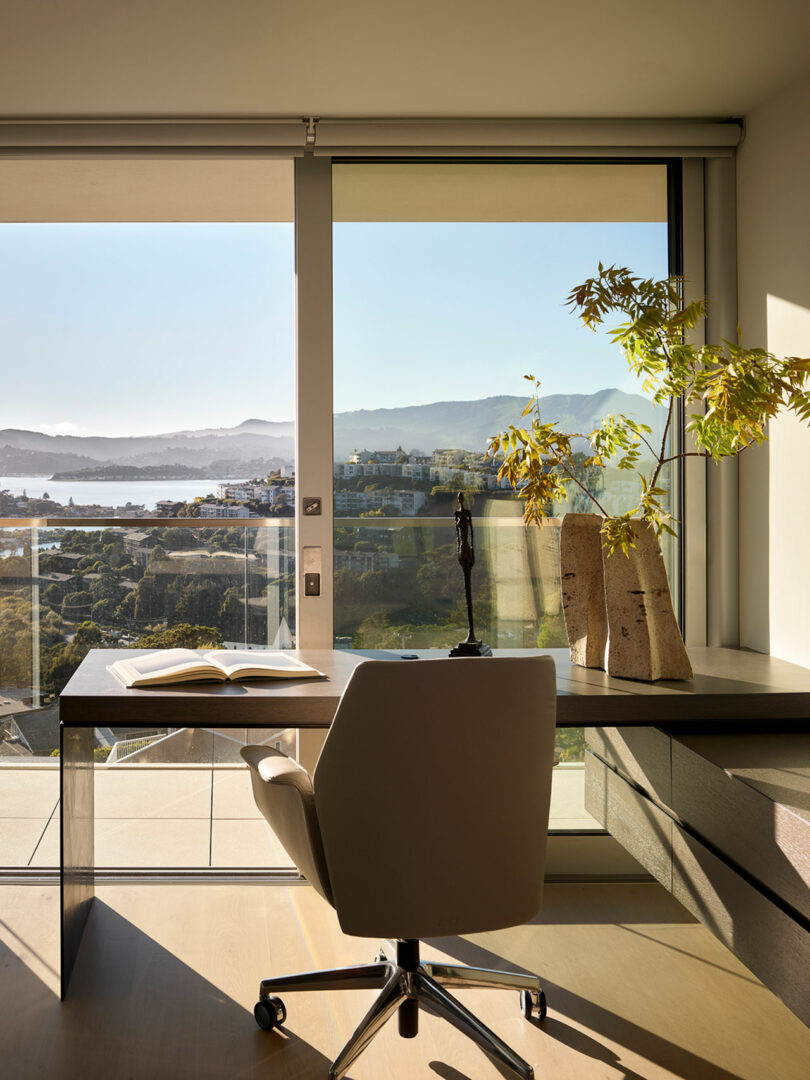 Modern home office with a chair, desk, open book, sculpture, and vases with greenery, set beside large windows overlooking a scenic landscape with hills and water.
