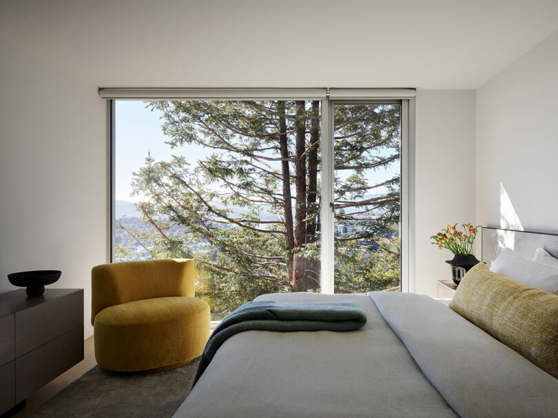 A modern bedroom with a large window showing a tree outside, featuring a yellow chair, gray dresser, and a neatly made bed with neutral bedding and yellow accents.
