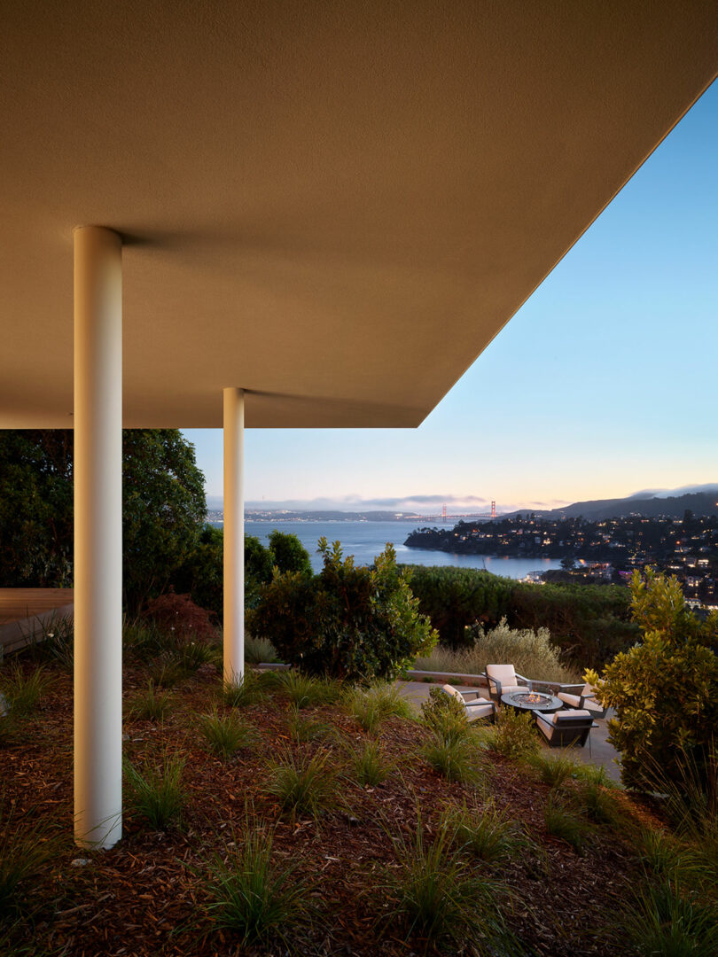 Modern patio with white columns and overhanging roof, overlooking a landscaped garden, water, and a distant cityscape at dusk.