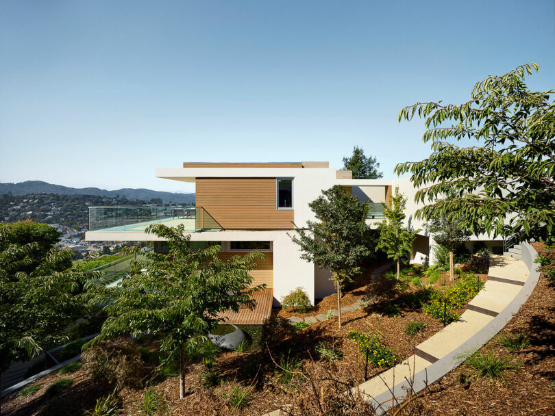 Modern house with wood and white exterior, surrounded by trees and landscaping, situated on a hillside with a view of distant mountains and city.
