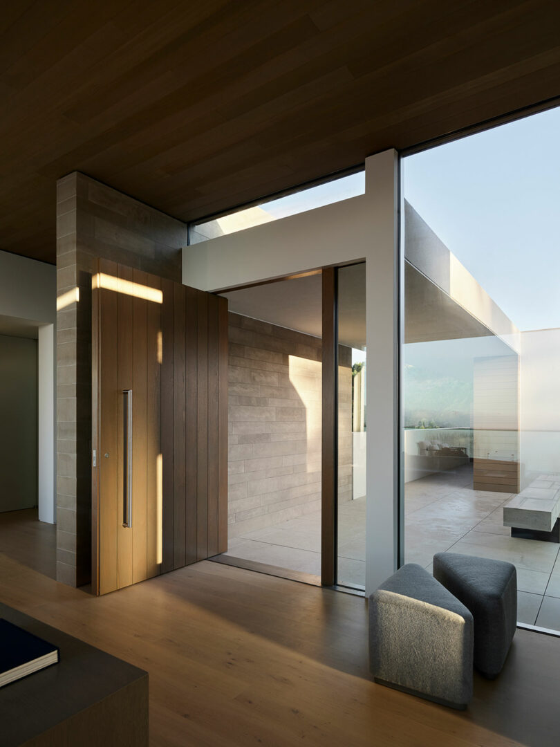 Modern minimalist entryway with large wooden pivot door, floor-to-ceiling glass panels, wood flooring, and outdoor seating area visible through the glass.