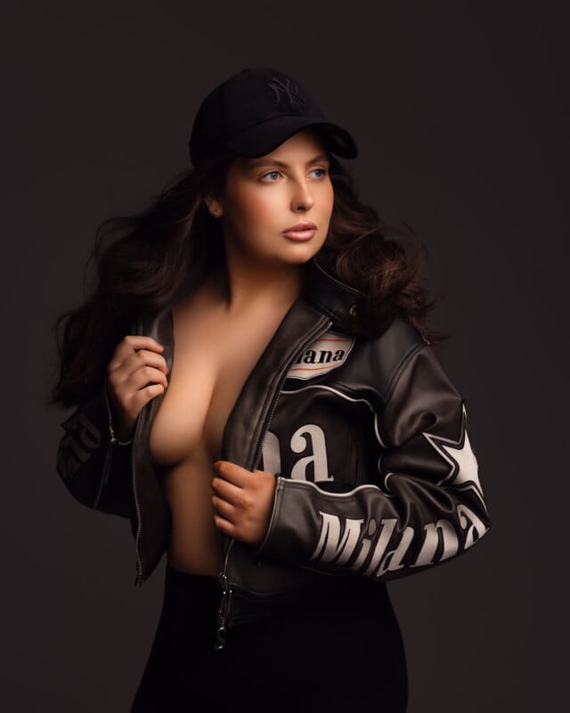 A woman with long brown hair wears a black baseball cap and a partially open black leather jacket, exposing her chest. She looks to the side against a dark background.
