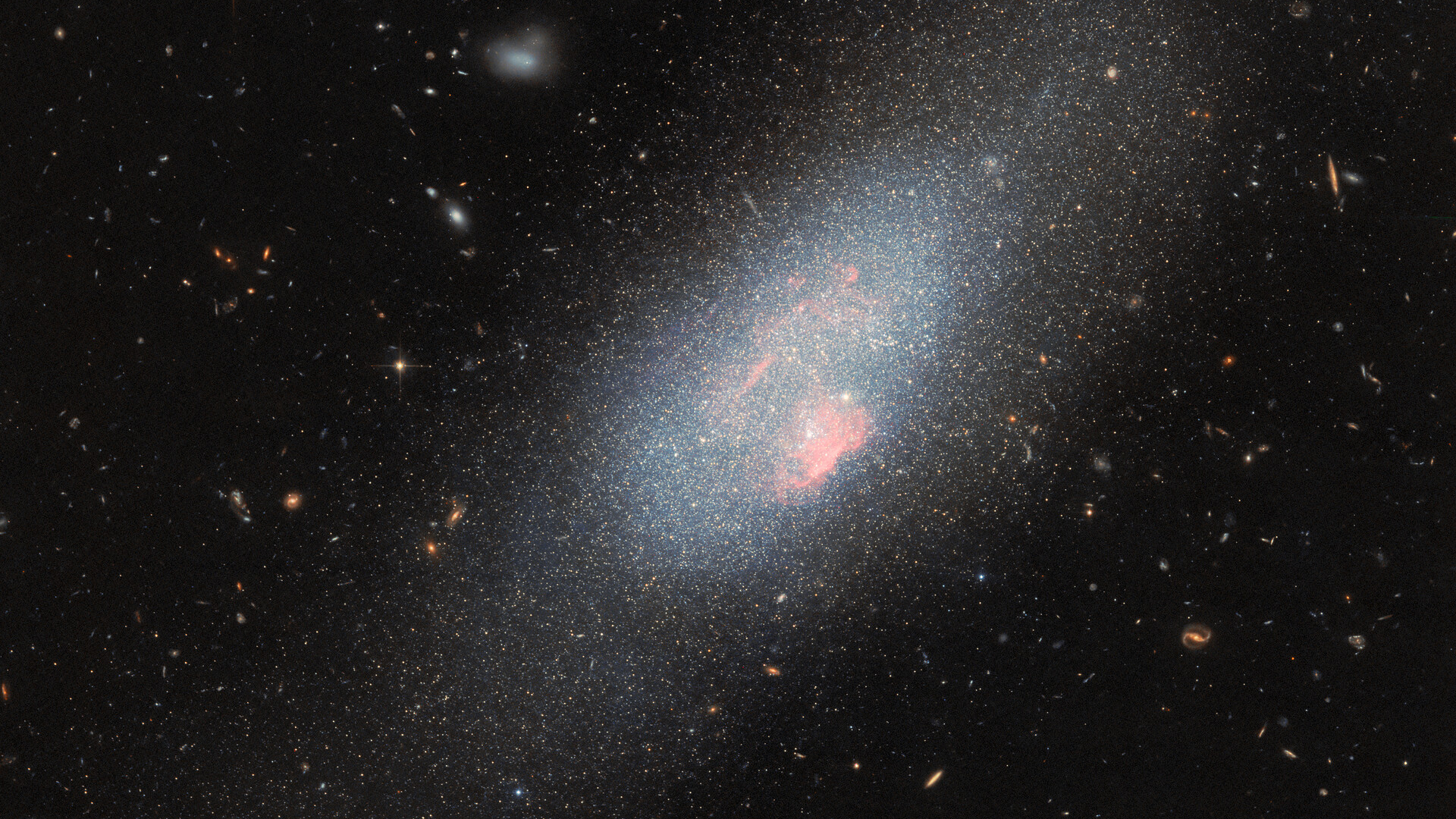 A glowing swirl of lights surrounds denser areas of pink color with stars surrounding the galaxy in a deep space image.