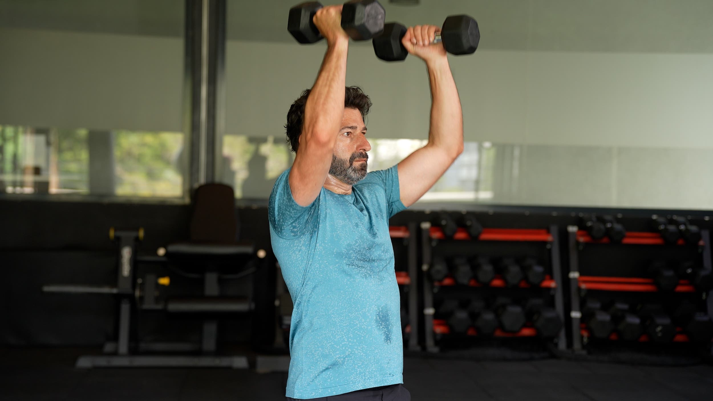 main with dark hair and a beard wearing a blue shirt presses two dumbbells above his shoulders
