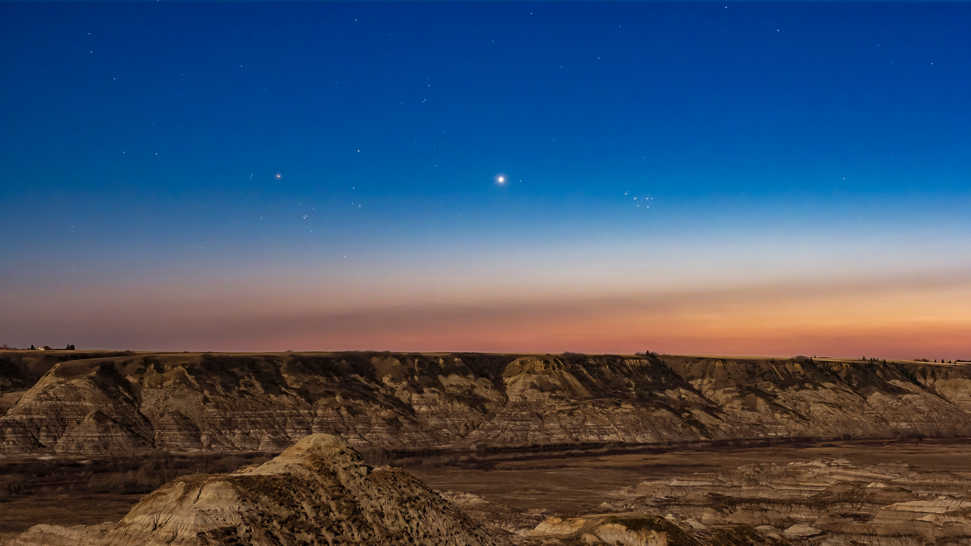 A deep blue night sky shines over a series of rocky plateaus while a bright dot, Venus, is seen in the center of the sky