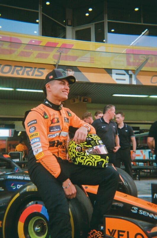 A race car driver in an orange suit and cap smiles while sitting on the front wheel of a race car, holding a yellow helmet. Several team members and a garage are visible in the background.