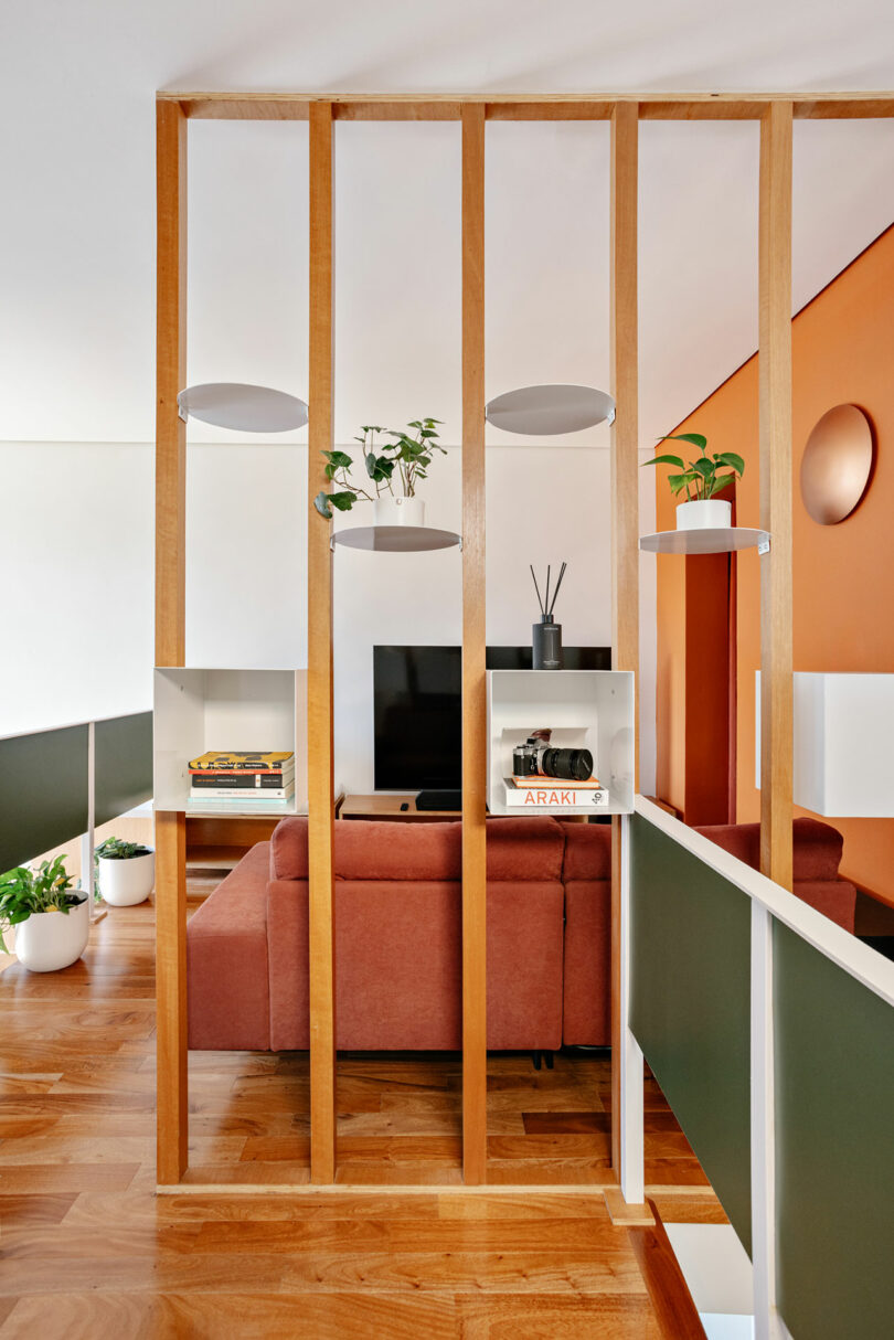 A modern living room with a wooden slat partition, shelves holding plants and books, a brown sofa, and an orange accent wall.