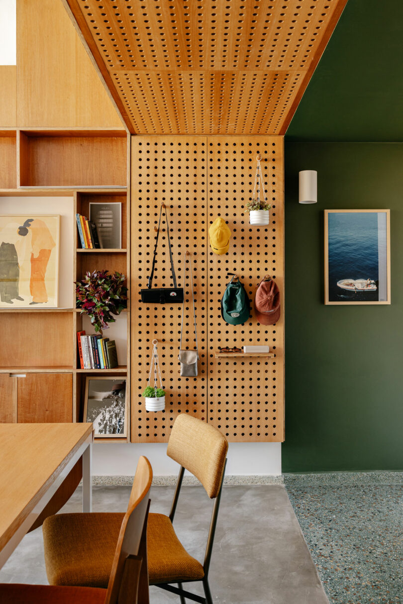 Modern interior with a wooden pegboard holding hats and bags, a bookshelf with books and art, and a green wall with framed artwork and a wall sconce.