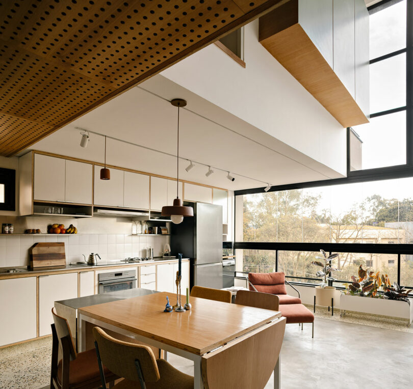 Modern kitchen and dining area with wooden furniture, large windows, built-in cabinets, pendant lights, and a view of trees outside.