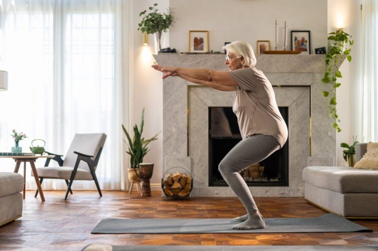 A senior woman performing a squat exercise on a yoga mat in a stylish, sunlit living room.