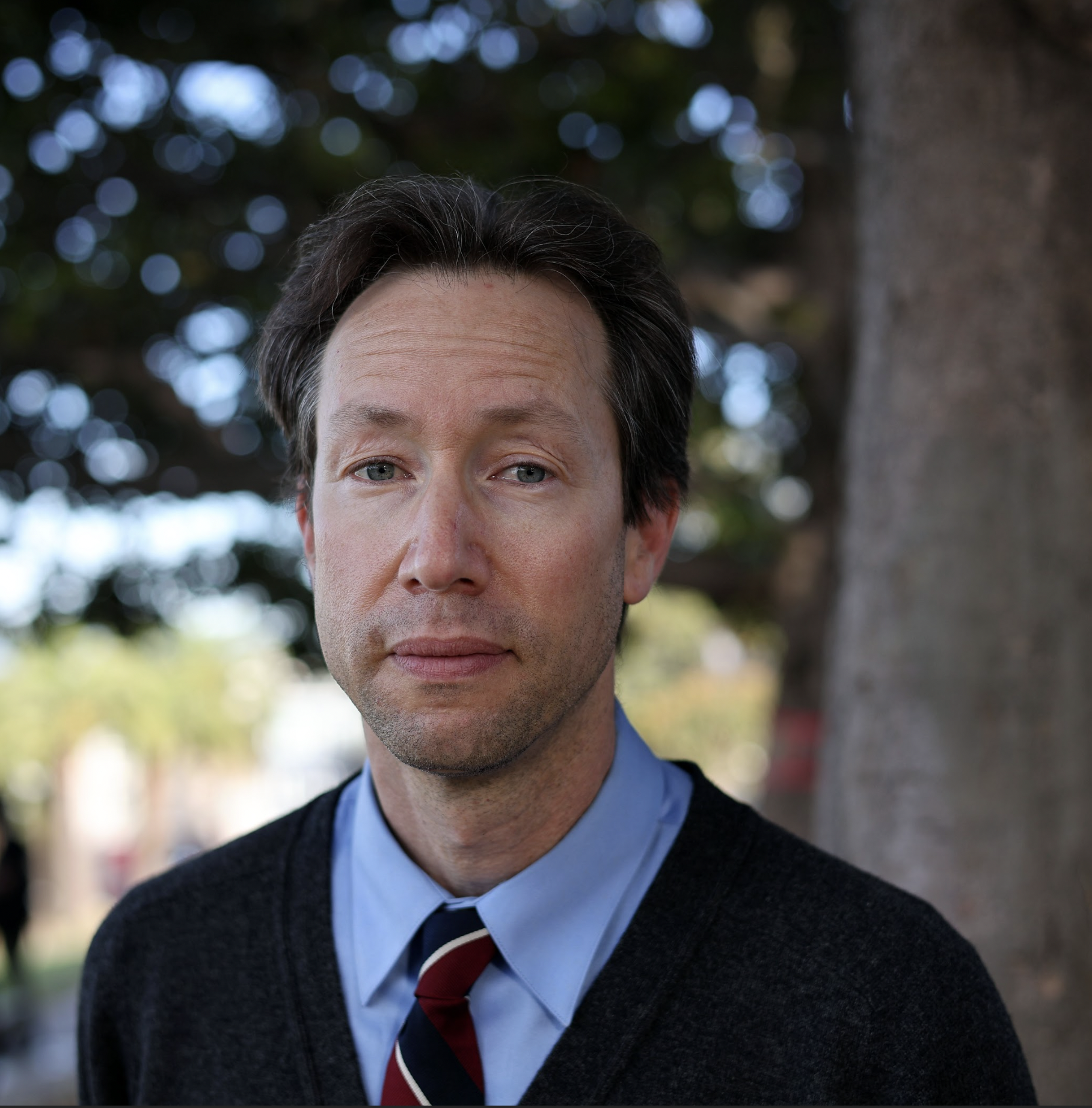 A person in a blue shirt and striped tie stands outdoors in front of a tree, looking at the camera.