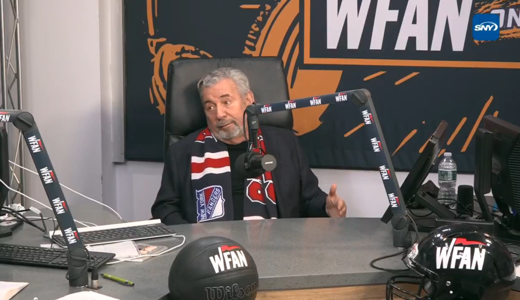 A man wearing a New York Rangers scarf sits at a desk with a WFAN microphone, a basketball, and a football helmet, all branded with WFAN.