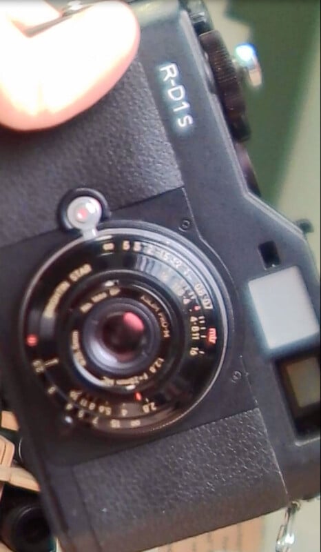 A close-up of a hand holding a black Epson R-D1s digital rangefinder camera, showing its lens, dials, buttons, and part of the grip.