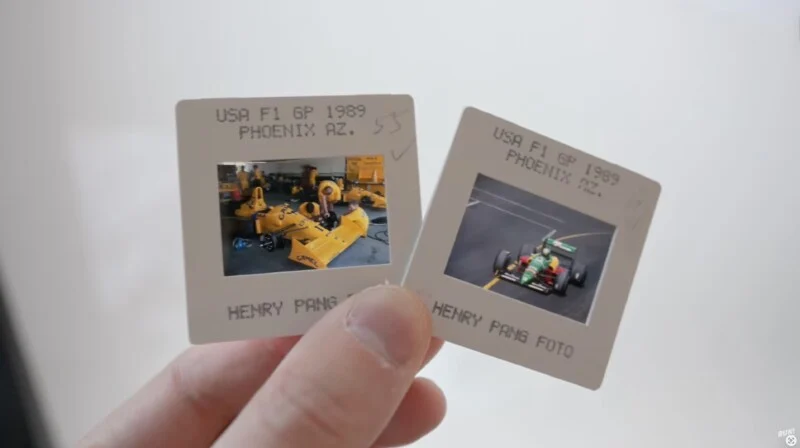 A hand holding two photo slides labeled "USA F1 GP 1989 Phoenix AZ." One slide shows a yellow Formula 1 car being serviced, and the other shows a race car on the track. Both slides are credited to Henry Pang.