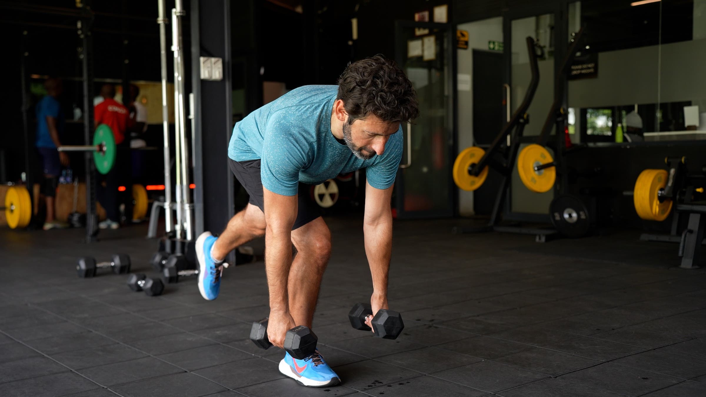 as part of a longevity workout plan, a man with dark hair wearing a blue shirt and black shorts demonstrates how to do a single-leg romanian deadlift (RDL)